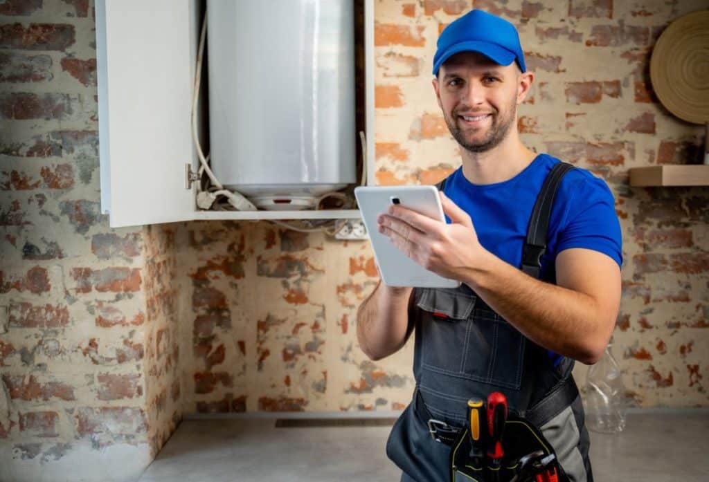 Plumber in blue workwear holding a tablet while servicing a residential water heater in a utility room with exposed brick walls.