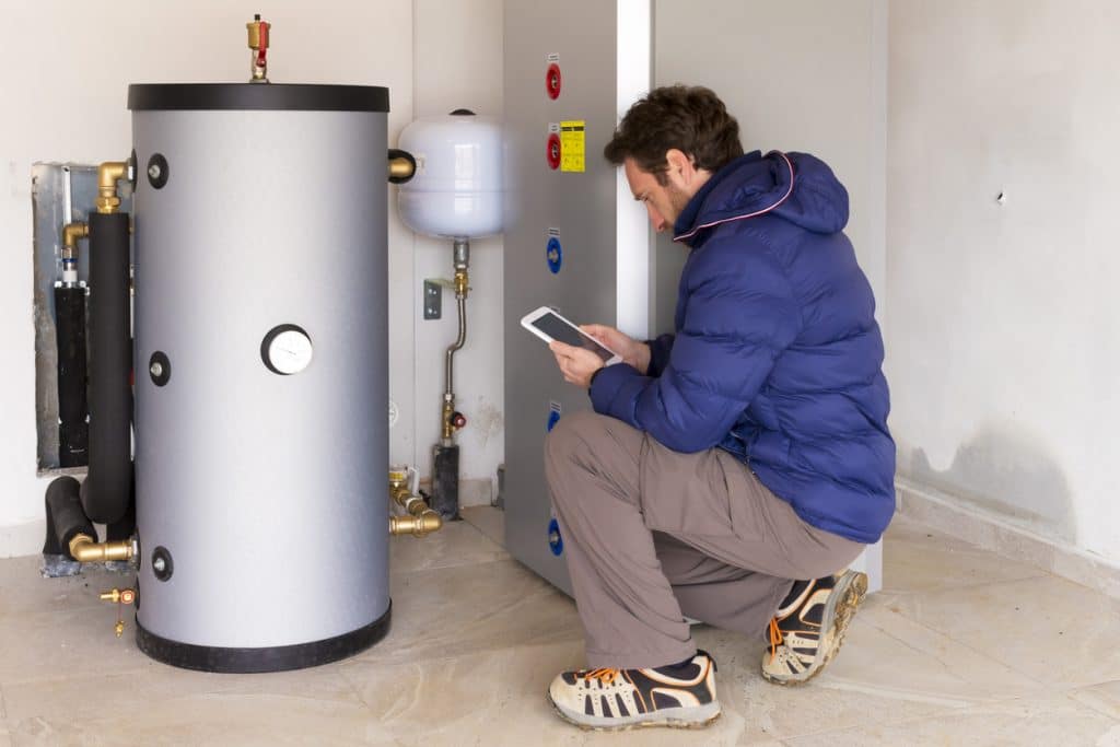 Technician inspecting a residential water heater system while reviewing information on a tablet beside the storage tank and connected plumbing.